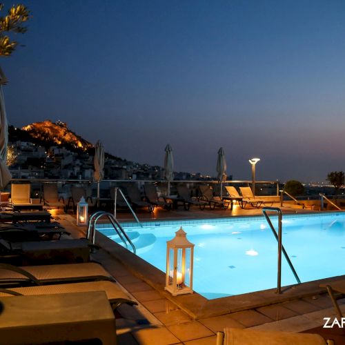Rooftop hotel pool at dusk with sun loungers, umbrellas, lanterns, and a city hillside view across the sky.