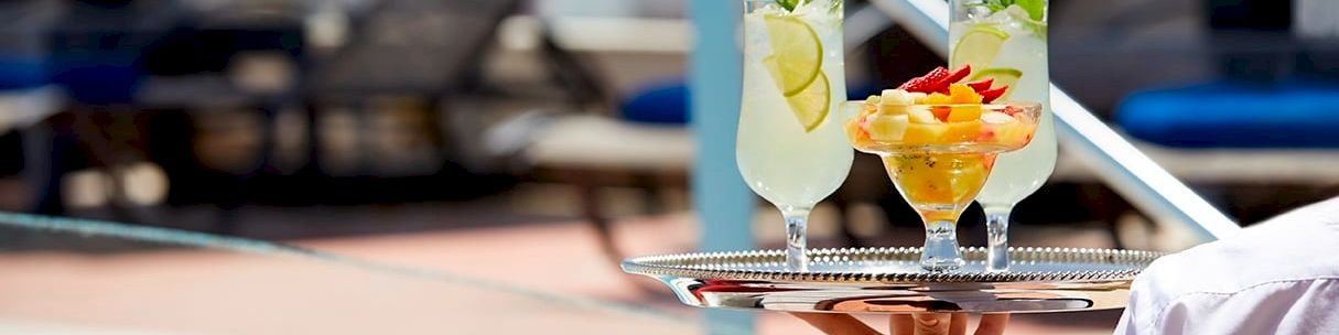 Two colorful cocktails with fruit garnish are served on a tray by a sunny pool, held by a waiter in white.