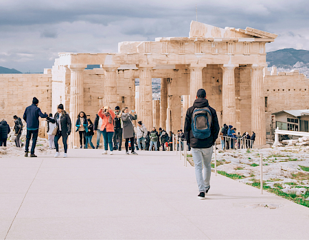 Tourists walk in front of the Parthenon at the Acropolis in Athens, Greece under a cloudy sky.