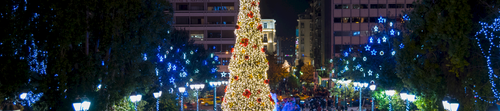 A huge lit Christmas tree with a glowing star tops a crowded city street, with blue light displays and fountains.