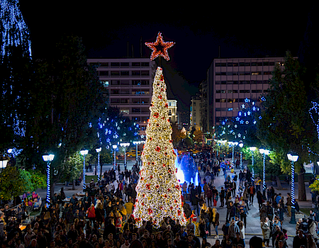 A huge lit Christmas tree with a glowing star tops a crowded city street, with blue light displays and fountains.