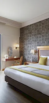 A modern hotel bedroom with a king bed, bedside lamps, and curtains by a glass door, plus a patterned accent wall.