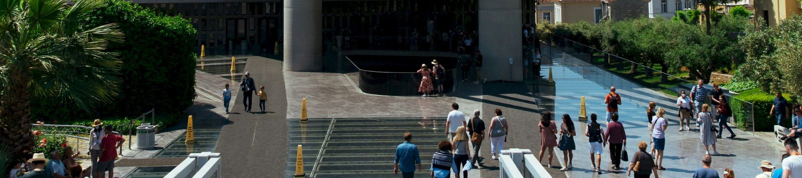 A crowd strolls through a modern outdoor museum plaza with a glass facade, ramps, and greenery under a striped canopy.